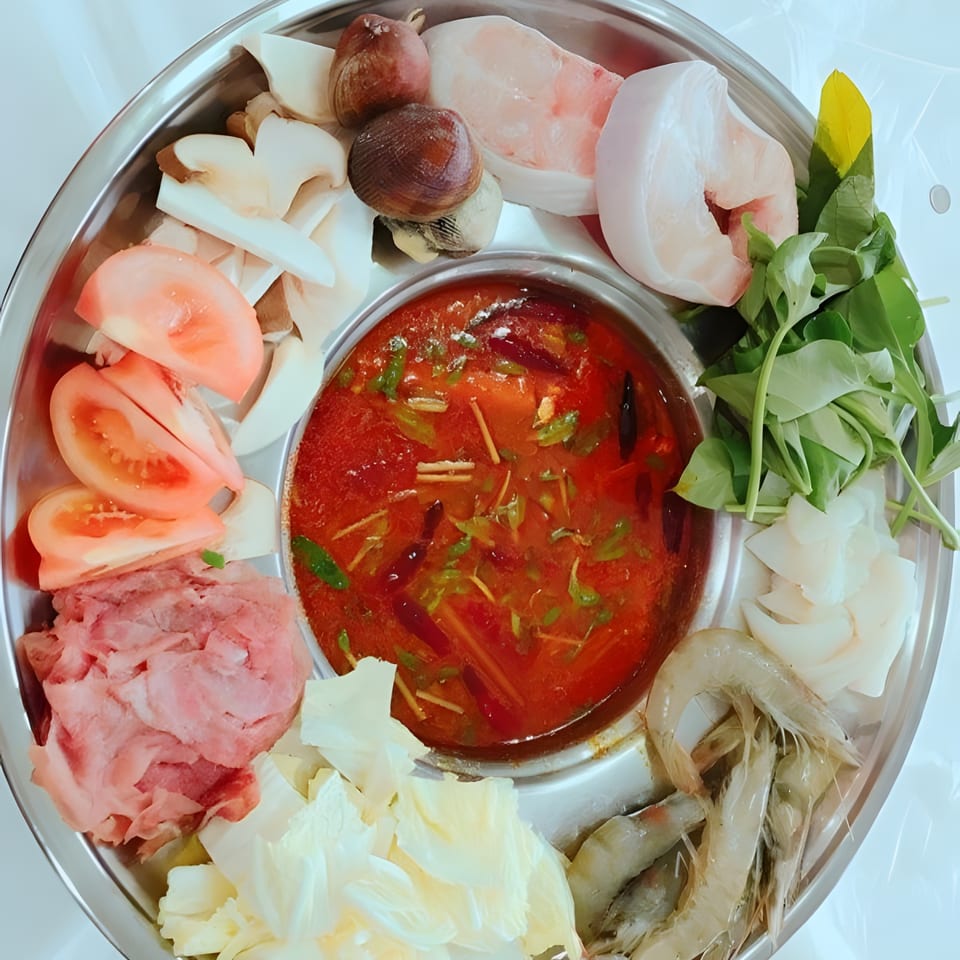 A hot pot with red broth surrounded by raw shrimp, meat, vegetables, and mushrooms.