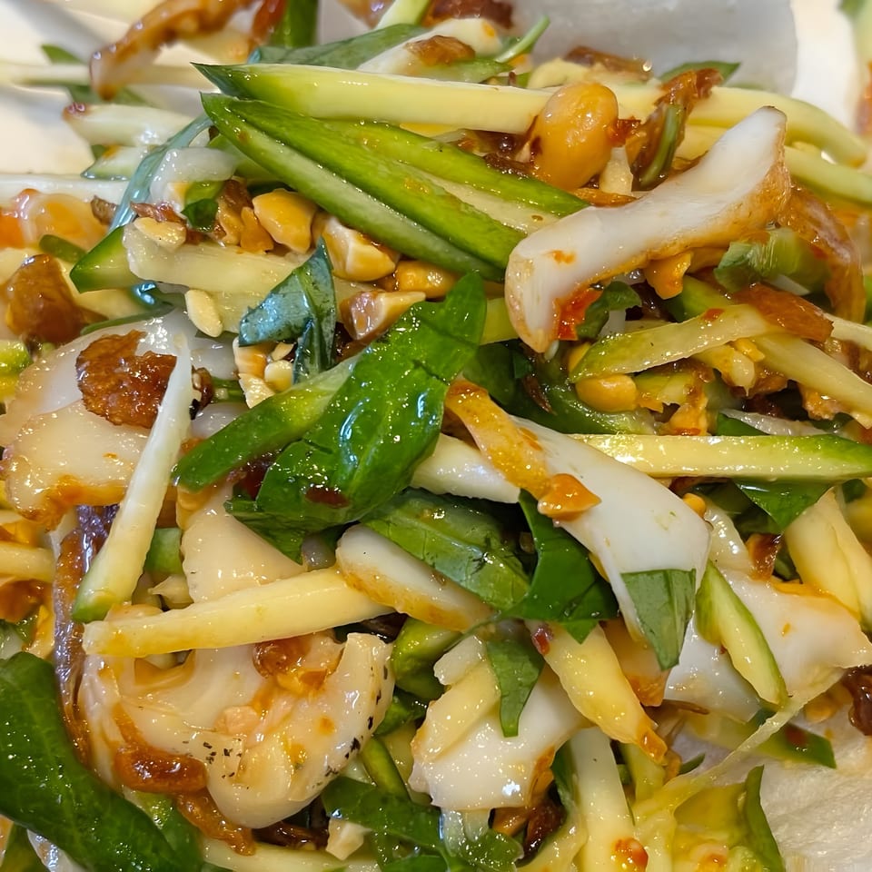 Close-up of a vibrant salad with green vegetables, white seafood, and peanuts on a white plate.