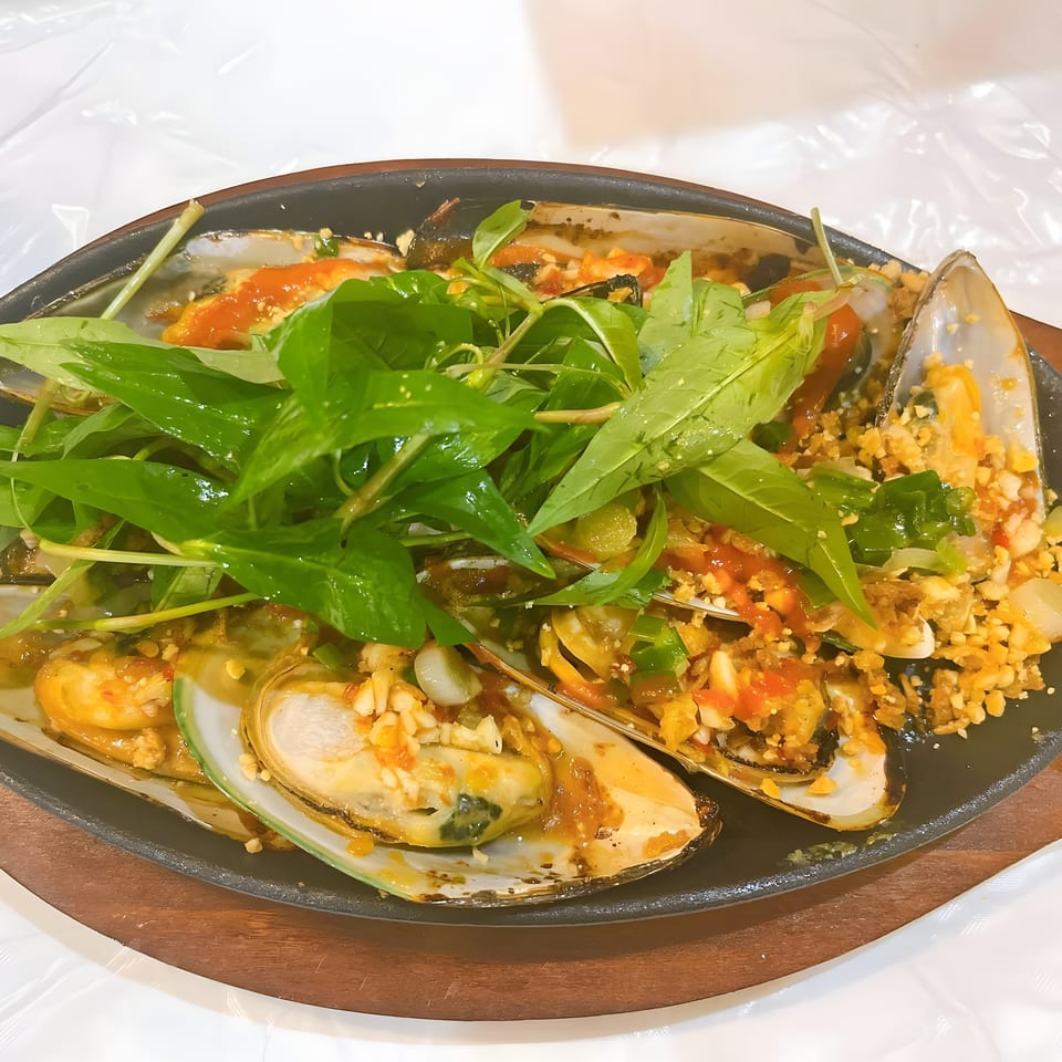 Sizzling mussels topped with green herbs, chili sauce, and crushed peanuts on a wooden platter.